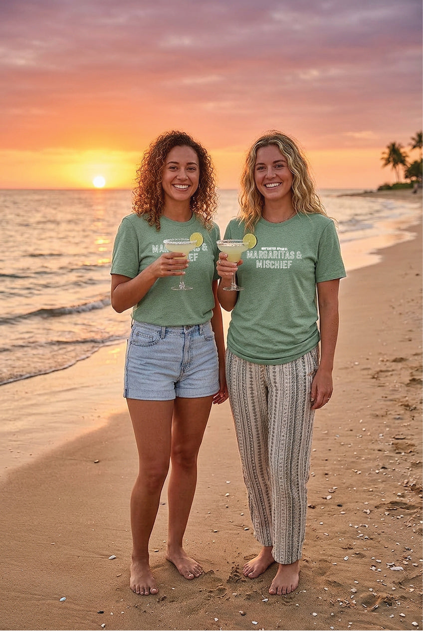 Two women standing on a beach at sunset, wearing green t-shirts with text, holding drinks.