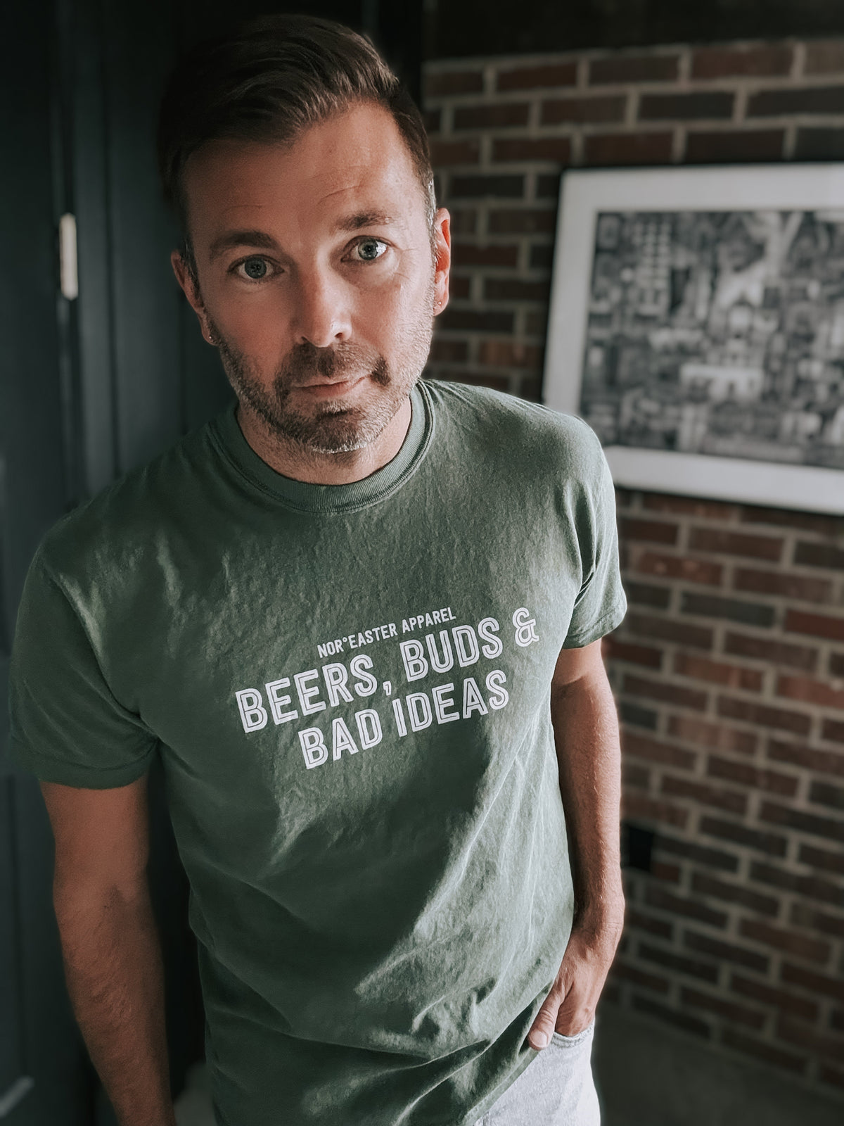 Man wearing a green t-shirt with text, standing in front of a brick wall.