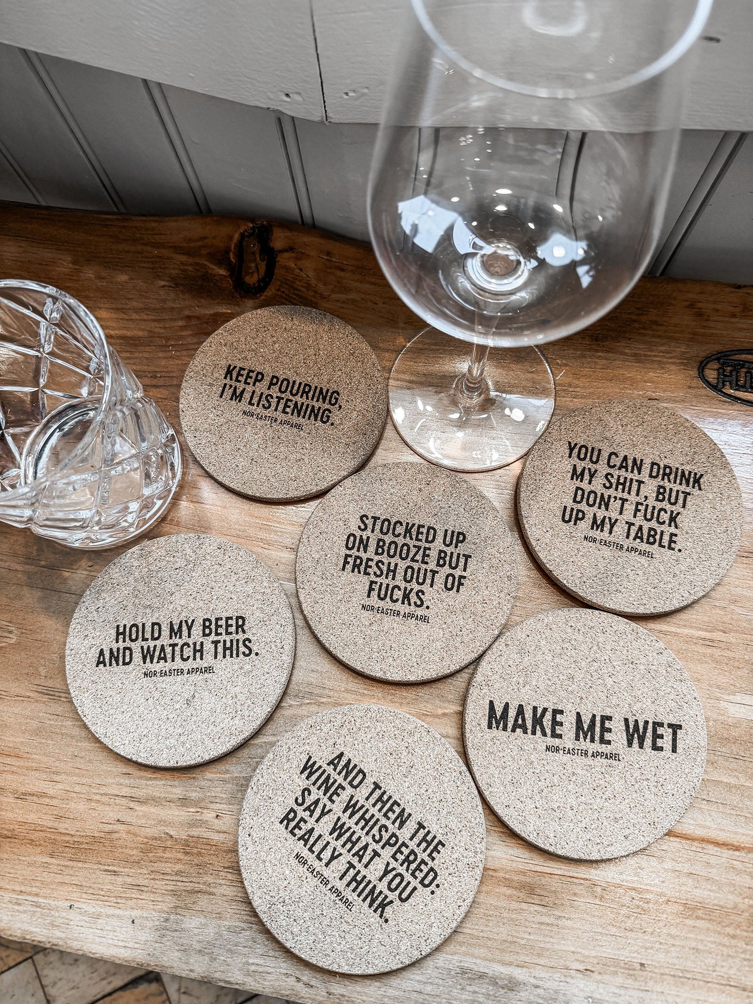 Set of humorous coasters with witty sayings on a wooden surface with wine glasses.