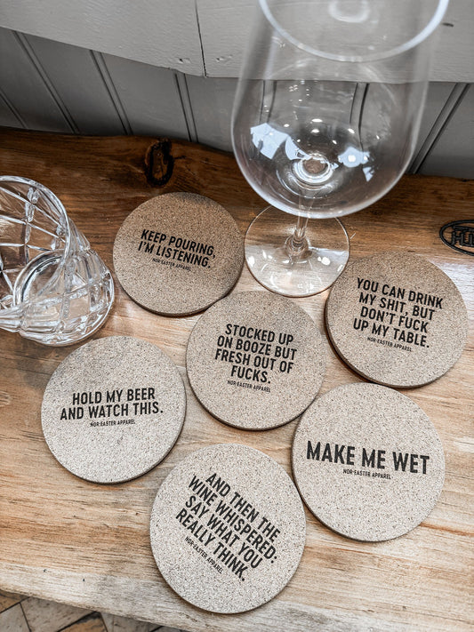 Set of humorous coasters with witty sayings on a wooden surface with wine glasses.