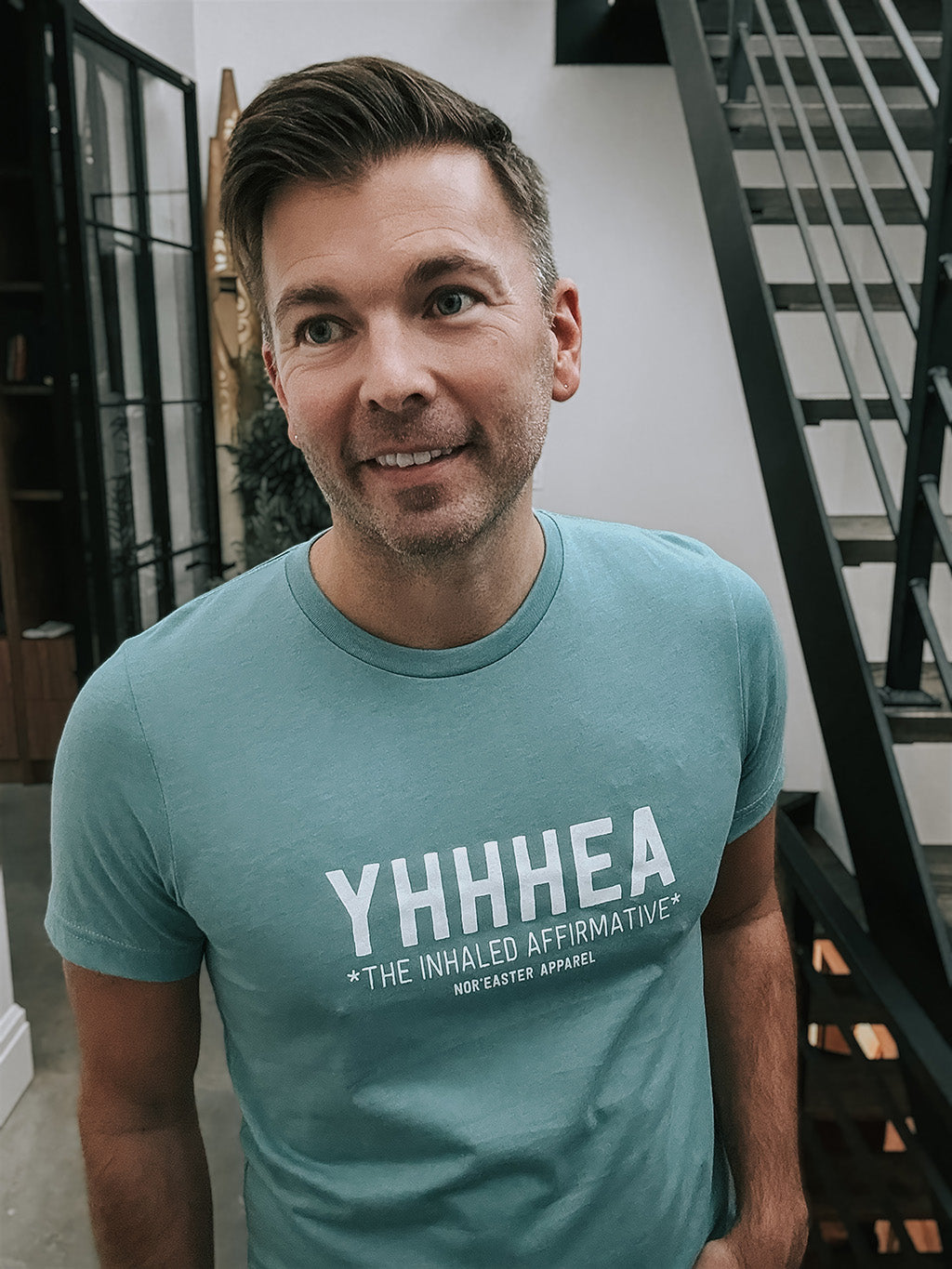 Man wearing a teal t-shirt with 'YHHHEA' text, standing indoors.